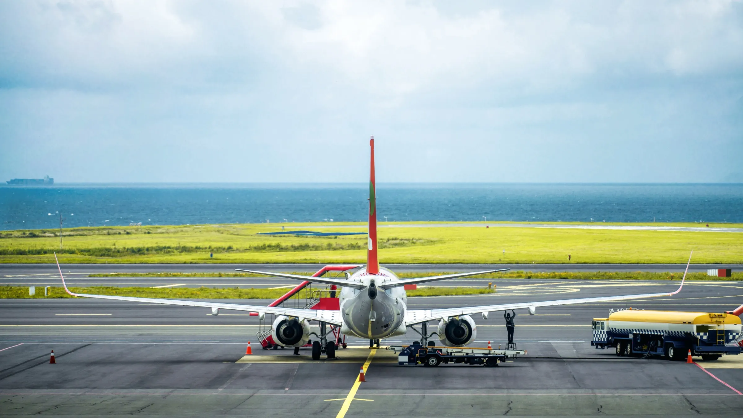 Un aereo di linea fermo sul piazzale di un aeroporto, con la pista e una striscia di mare sullo sfondo, virato in blu